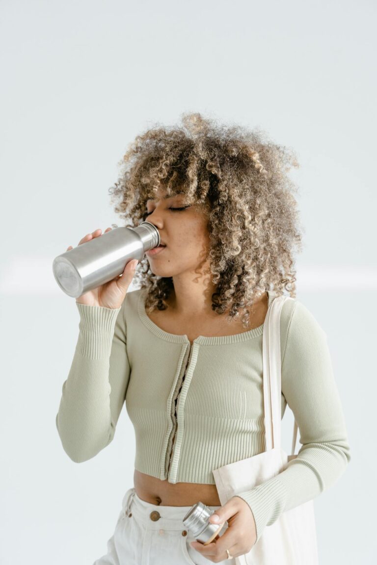 Stylish young woman with curly hair drinking water from a reusable stainless steel bottle.